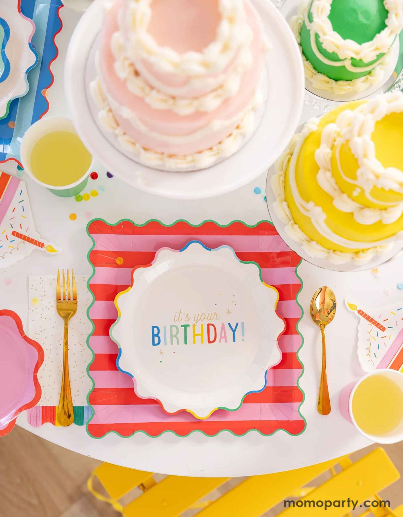 A festive and cheerful birthday party table featuring Momopartys's Happy Birthday party tablewares including the 9" It's Your Birthday Plates, the red striped dinner plates, the slice of cake shaped napkins, and the rainbow fringe guest towels by My Mind's Eye. On the table there are multiple birthday cakes in pastel rainbow colors, making this a modern and adorable table setting for a happy birthday celebration for kids of any age!