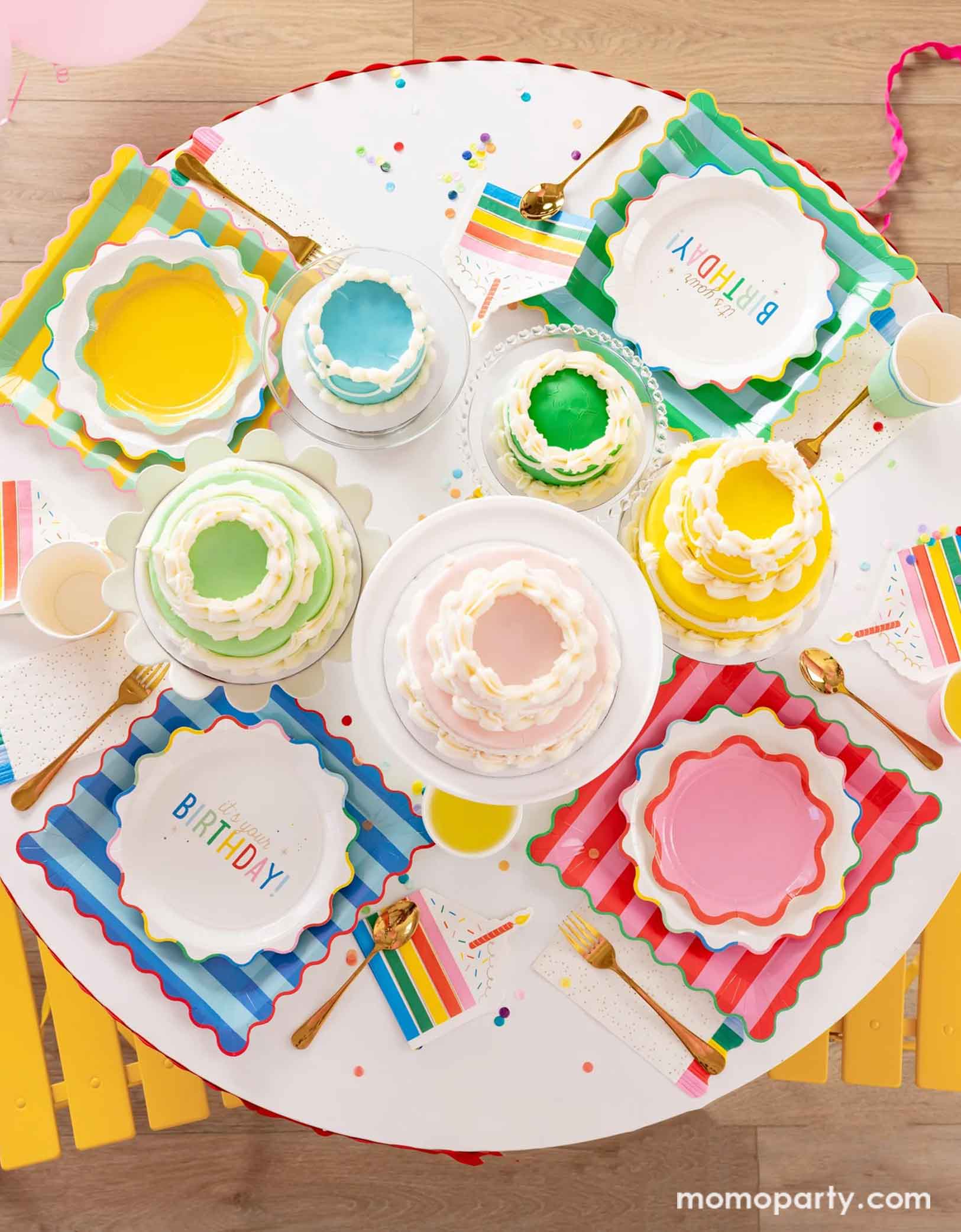 A overhead shot of a festive and cheerful round birthday party table featuring Momopartys's Happy Birthday party tablewares including the 9" It's Your Birthday Plates, the striped dinner plates in four different colors the ric rac edge dessert plates, the slice of cake shaped napkins, and the rainbow fringe guest towels. On the table there are multiple birthday cakes in pastel rainbow colors in different sizes, making this a modern table setting for a happy birthday celebration for kids of any age!