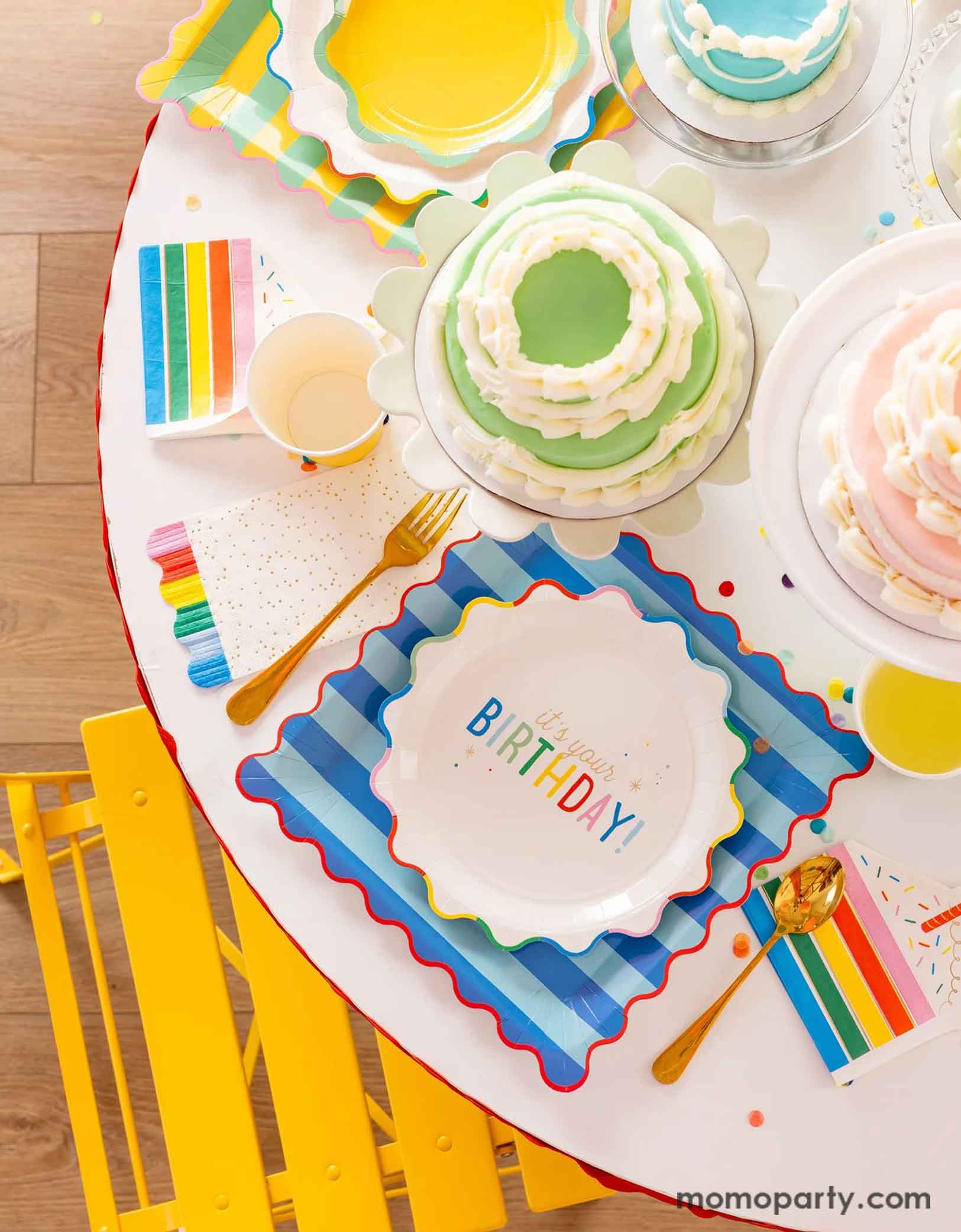 A festive and cheerful birthday party table featuring Momopartys's Happy Birthday party tablewares including the 9" It's Your Birthday Plates, the striped dinner plates, the slice of cake shaped napkins, the rainbow fringe guest towels by My Mind's Eye. On the table there are multiple birthday cakes in pastel rainbow colors, making this a modern and adorable table setting for a happy birthday celebration for kids of any age!