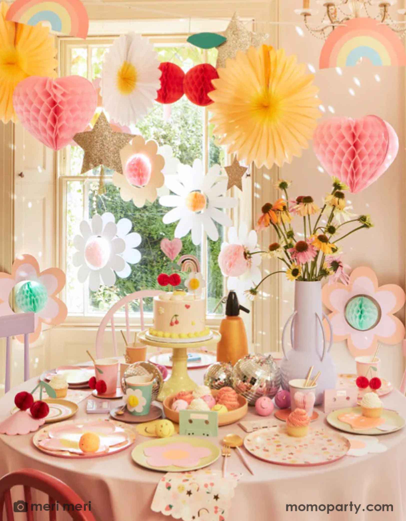 A 90's inspired party table featuring Momopartys's Happy Face Icons party collection by Meri Meri, including happy smiley flower shaped plates, happy icon dinner plates, large napkins and party bags and die-cut napkins in rainbow, smiley face and mushroom shape. Above the table there's a garland with 3D honeycomb decorations of hearts, daisies, rainbows, giving this table a nostalgic and groovy vibe. Perfect for any happy occasion!