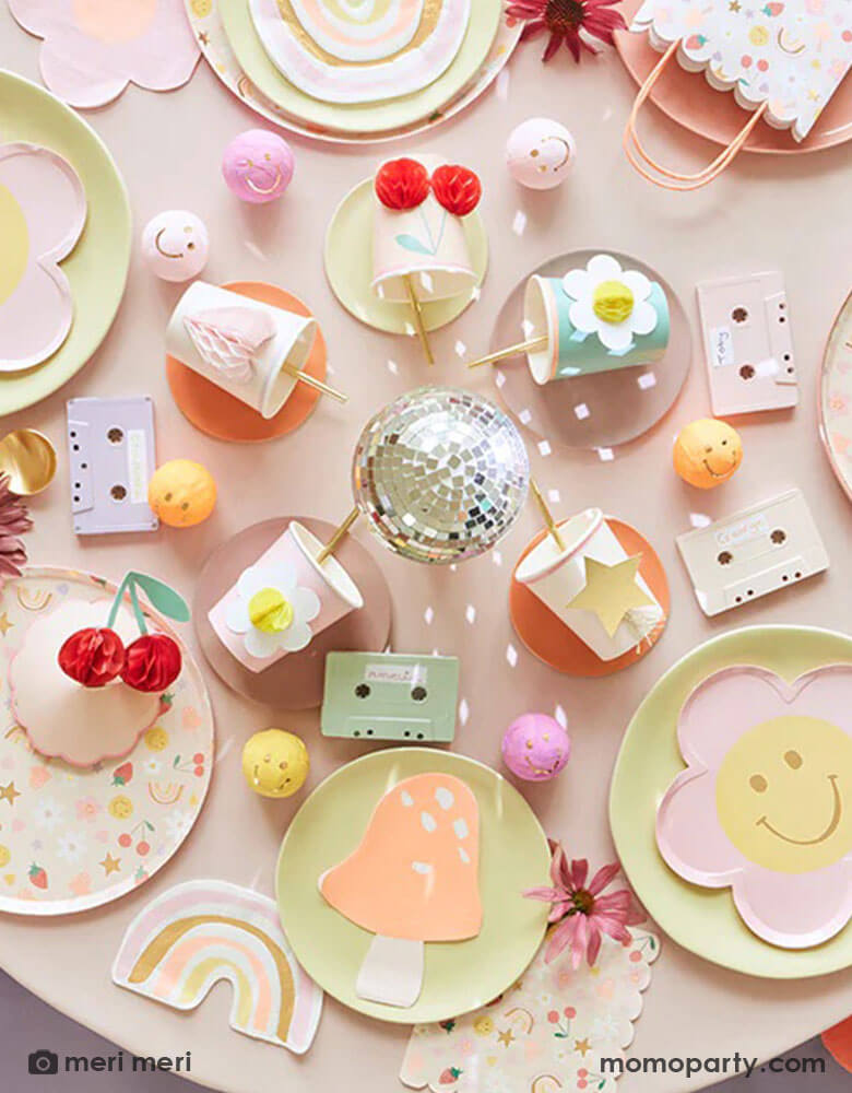 A 90's inspired party table featuring Momopartys's Happy Face Icons party collection by Meri Meri, including happy smiley flower shaped plates, happy icon dinner plates, large napkins and party bags and die-cut napkins in rainbow, smiley face and mushroom shape. In the middle there's a disco ball and some cassette tapes as the centerpiece which are surrounded by Meri Meri's 3D honeycomb cups, giving this table a nostalgic and groovy vibe. Perfect for any happy occasion!