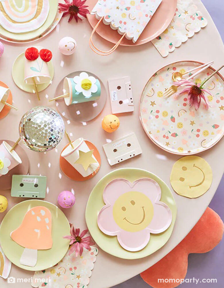 A 90's inspired party table featuring Momopartys's Happy Face Icons party collection by Meri Meri, including happy smiley flower shaped plates, happy icon dinner plates, large napkins and party bags and die-cut napkins in rainbow, smiley face and mushroom shape. In the middle there's a disco ball and some cassette tapes as the centerpiece which are surrounded by Meri Meri's 3D honeycomb cups, giving this table a nostalgic and groovy vibe. Perfect for any happy occasion!