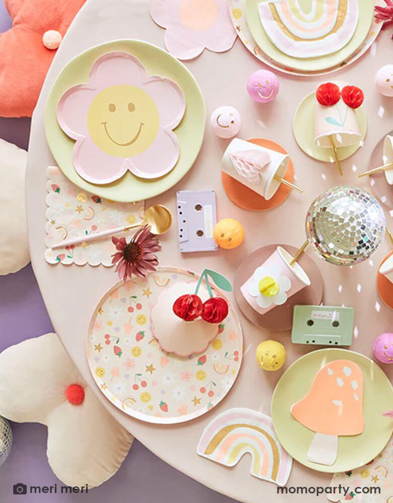 A 90's inspired party table featuring Momopartys's Happy Face Icons party collection by Meri Meri, including happy smiley flower shaped plates, happy icon dinner plates, large napkins and party bags and die-cut napkins in rainbow, smiley face and mushroom shape. In the middle there's a disco ball and some cassette tapes as the centerpiece which are surrounded by Meri Meri's 3D honeycomb cups, giving this table a nostalgic and groovy vibe. Perfect for any happy occasion!