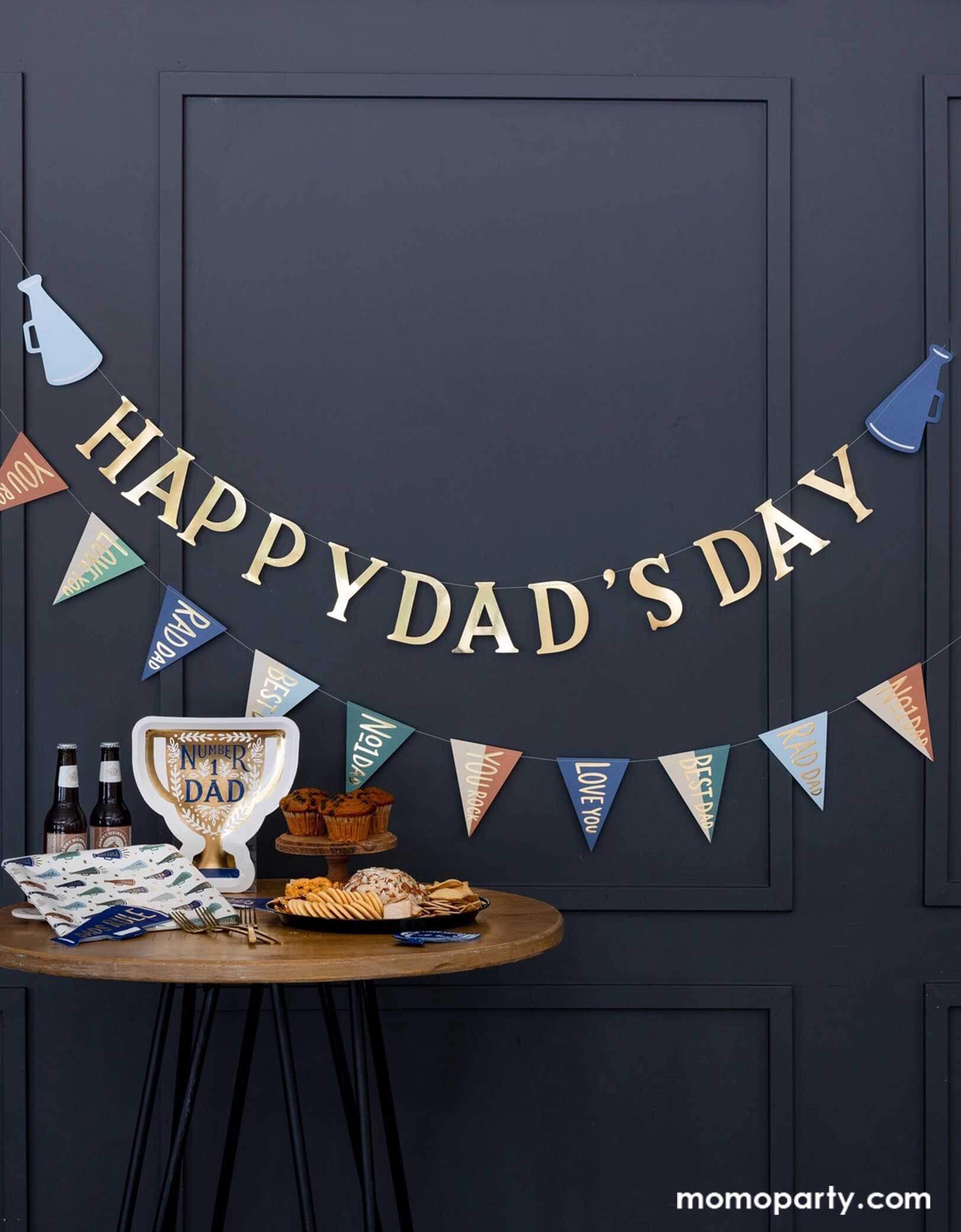 A navy room decorated with Momopartys's Father's Day party decors by My Mind's Eye including the 6' Father's Day Banner Set hung on the wall, which one banner says Happy Dad's Day, the other has pennant flags. One the table, there are some cupcakes, a charcuterie board and some beer bottled beverages. Along with party tableware including Number One Dad trophy shaped plates, megaphone pattern square dinner plates and Dads Rule napkins, this set up is perfect to celebrate dads!