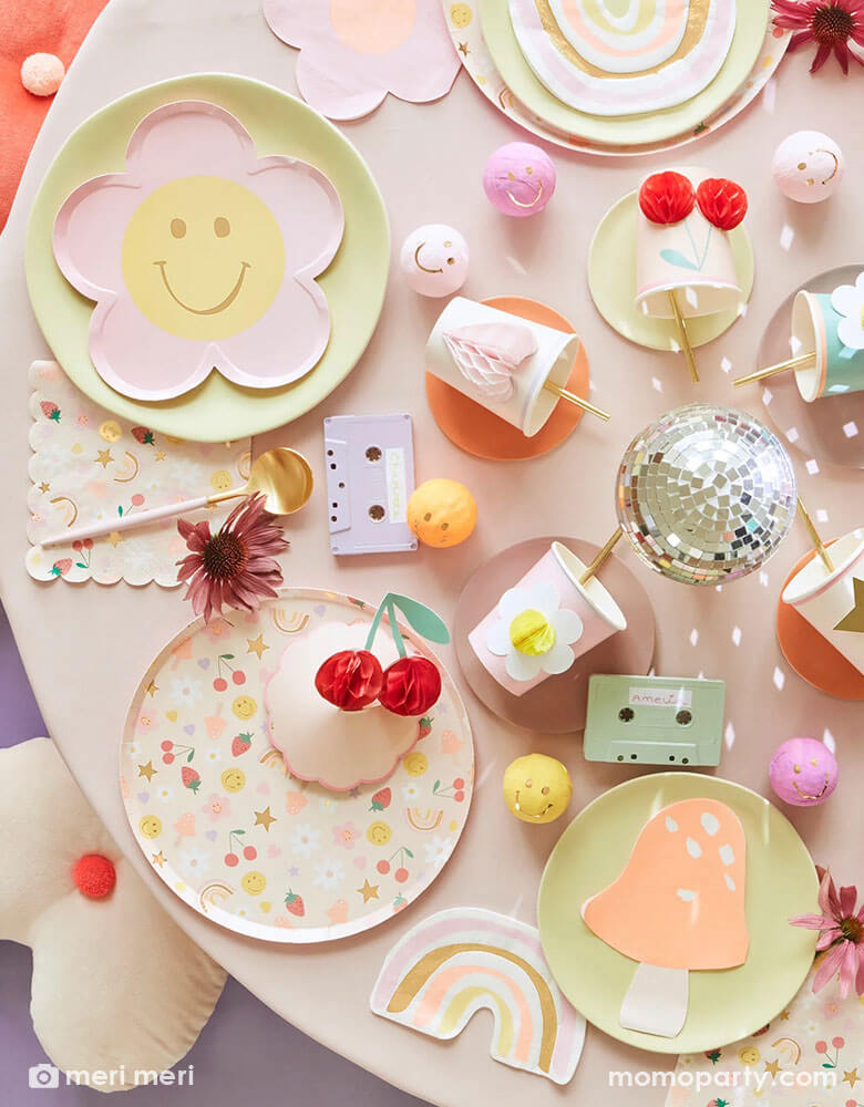 A 90's inspired party table featuring Momopartys's Happy Face Icons party collection by Meri Meri, including happy smiley flower shaped plates, happy icon dinner plates, large napkins and party bags and die-cut napkins in rainbow, smiley face and mushroom shape. In the middle there's a disco ball and some cassette tapes as the centerpiece which are surrounded by Meri Meri's 3D honeycomb cups, giving this table a nostalgic and groovy vibe. Perfect for any happy occasion!