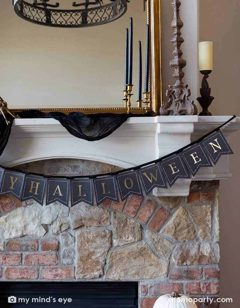 A close up shot of Momopartys's Happy Halloween banner hung over a mantel that's decorated with elegant Halloween decorations including a black and gold chandelier, black and gold candle holders, a beige colored pumpkin and a gold framed vintage mirror.