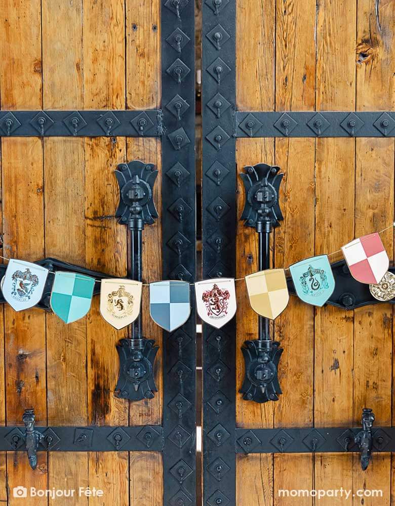 A big Harry Potter Hogwarts-like castle gate adorned with Momopartys's Harry Potter school pride multicolor party banner by Bonjour Fete featuring different Harry Potter house symbols and colors including Hufflepuff, Slytherin, Gryffindor and Ravenclaw - a perfect way to set the scene for a magical Harry Potter themed celebration, be it a kid's birthday party or a Halloween gathering.