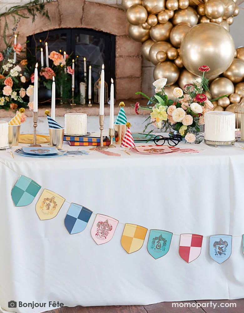 A Harry Potter themed party table adorned with Momopartys's Harry Potter school pride multicolor party banner featuring different Harry Potter house symbols and colors including Hufflepuff, Slytherin, Gryffindor and Ravenclaw. On the party table there are Harry Potter themed party tableware including plates, napkins, party hats, gold cups and white and gold candle sticks. In the back of the party table is a old vintage fireplace adorned with flowers and an organic gold balloon garland inspired by Quidditch.