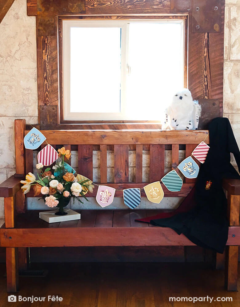 An old, medieval-like wooden bench adorned with Momopartys's Harry Potter school pride multicolor party banner featuring different Harry Potter house symbols and colors including Hufflepuff, Slytherin, Gryffindor and Ravenclaw. On the bench there's a white owl stuffy and a black Harry Potter robe hung across. With a beautiful flower arrangement, it makes a perfect idea for a welcome station that sets the scene for a Harry Potter themed celebration, be it a kid's birthday party or a Halloween gathering.