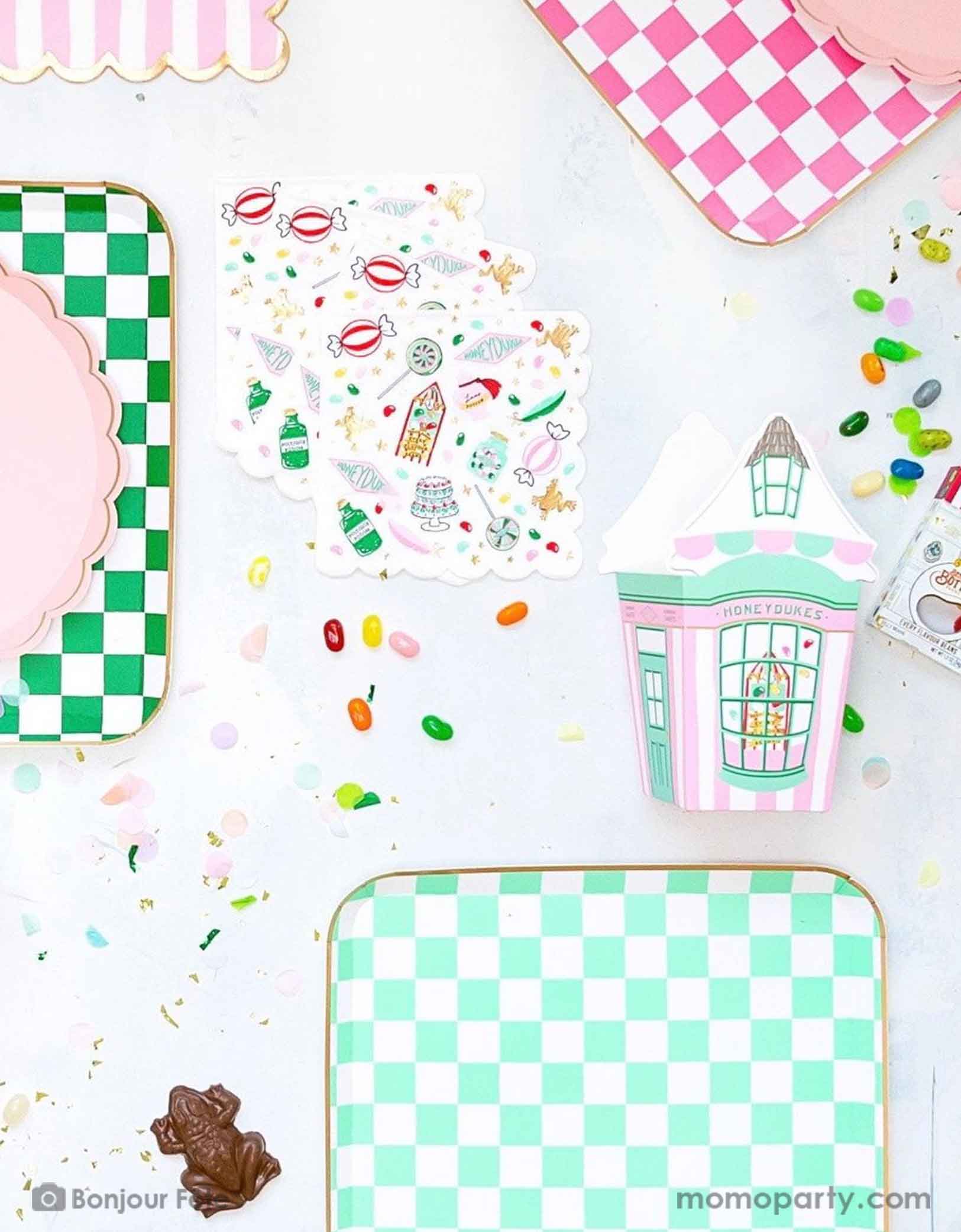 A Harry Potter themed tablescape featuring Momopartys's Harry Potter party collection including pink and green checkered dinner plates, pink striped Honeyduke large napkins, party cups and treat boxes. With chocolate frogs, Bertie Bott's Every Flavour Beans, lollipops and colorful confetti, this makes a festive scene for kid's Harry Potter themed birthday celebration.