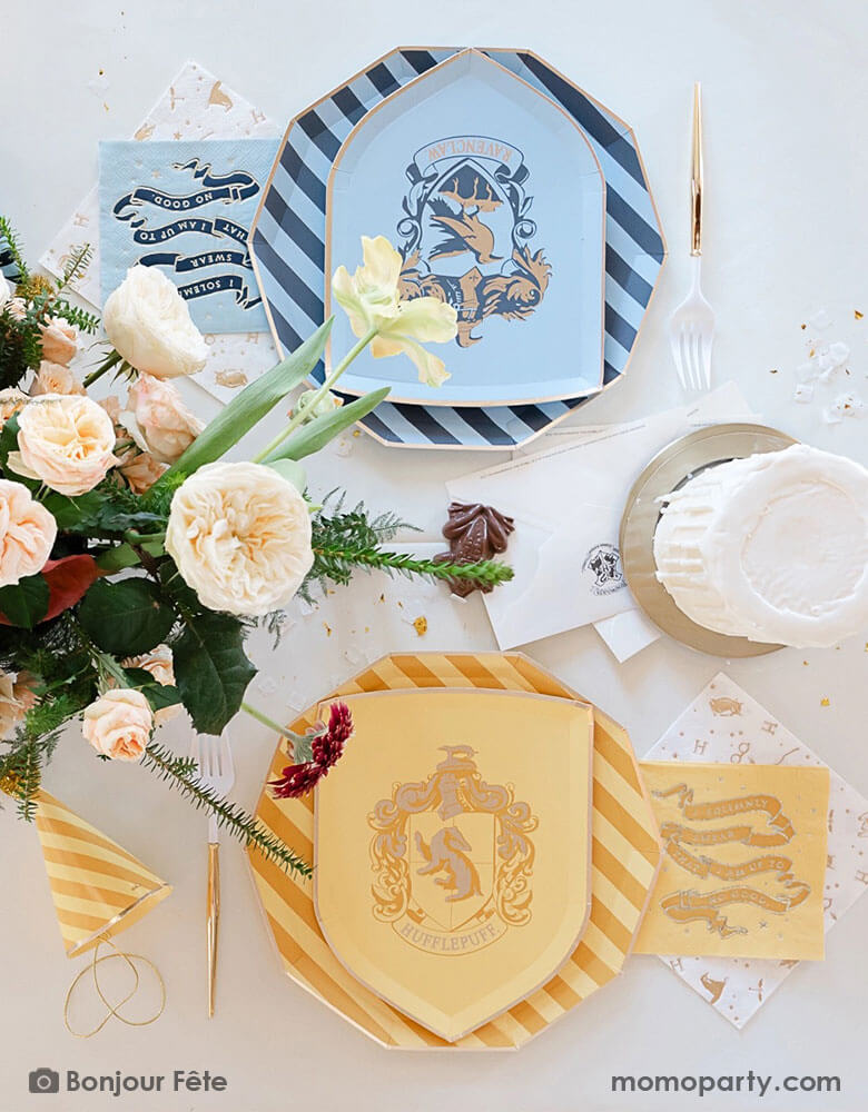 A beautiful and elegant Harry Potter themed party tablescape features Momopartys's Harry Potter house pride shield-shaped 7" side plates with Hufflepuff and Ravenclaw logos and colors paired with 10.25 striped dinner plates. Next to the plates are Harry Potter motif large napkins in gold foil and matching small napkins. On the table there's a candle stick shaped buttercream cake, a letter from Hogwarts, some gold confetti and party hats, along with a beautiful flower bouquet.