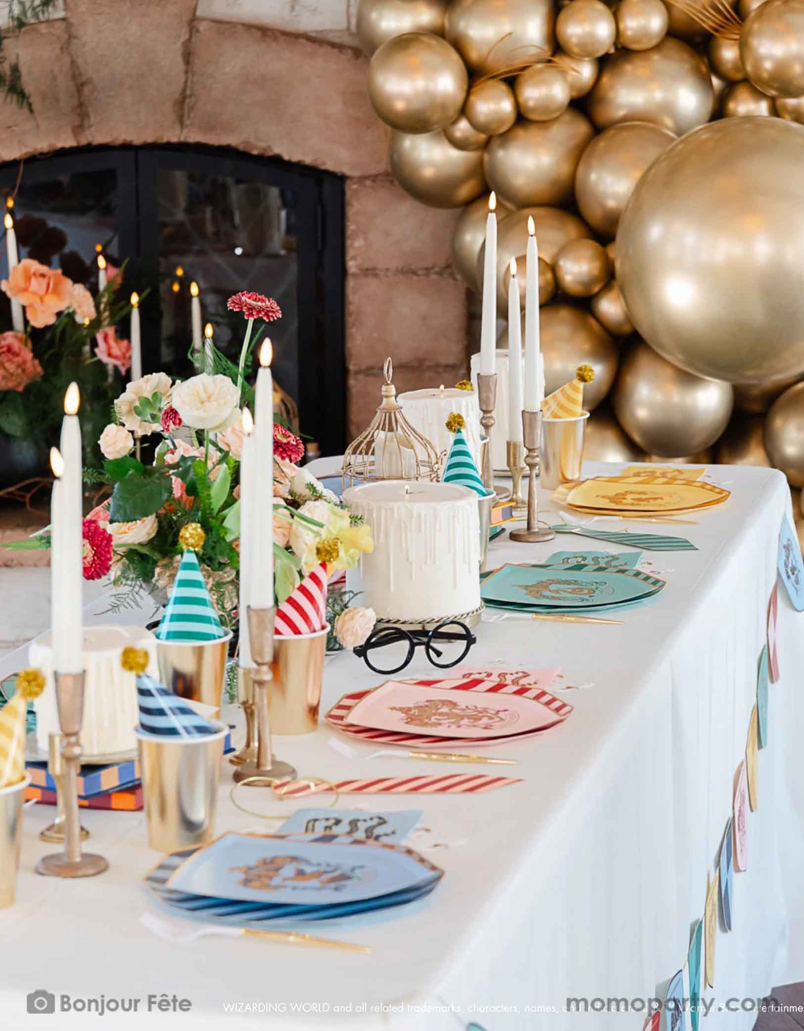 A beautiful Harry Potter themed party table setting featuring Momopartys's Harry Potter house pride shield-shaped 7" side plates with house logos and colors paired with 10.25 striped dinner plates and birthday party hats. In the center of the table there are some candle shaped birthday cakes, bird cages for owls, and some floral arrangement, plus a pair of Harry Potter's glasses. In the back there's a fire placed decorated with chrome gold balloon garland, making this a magical party for kids! 