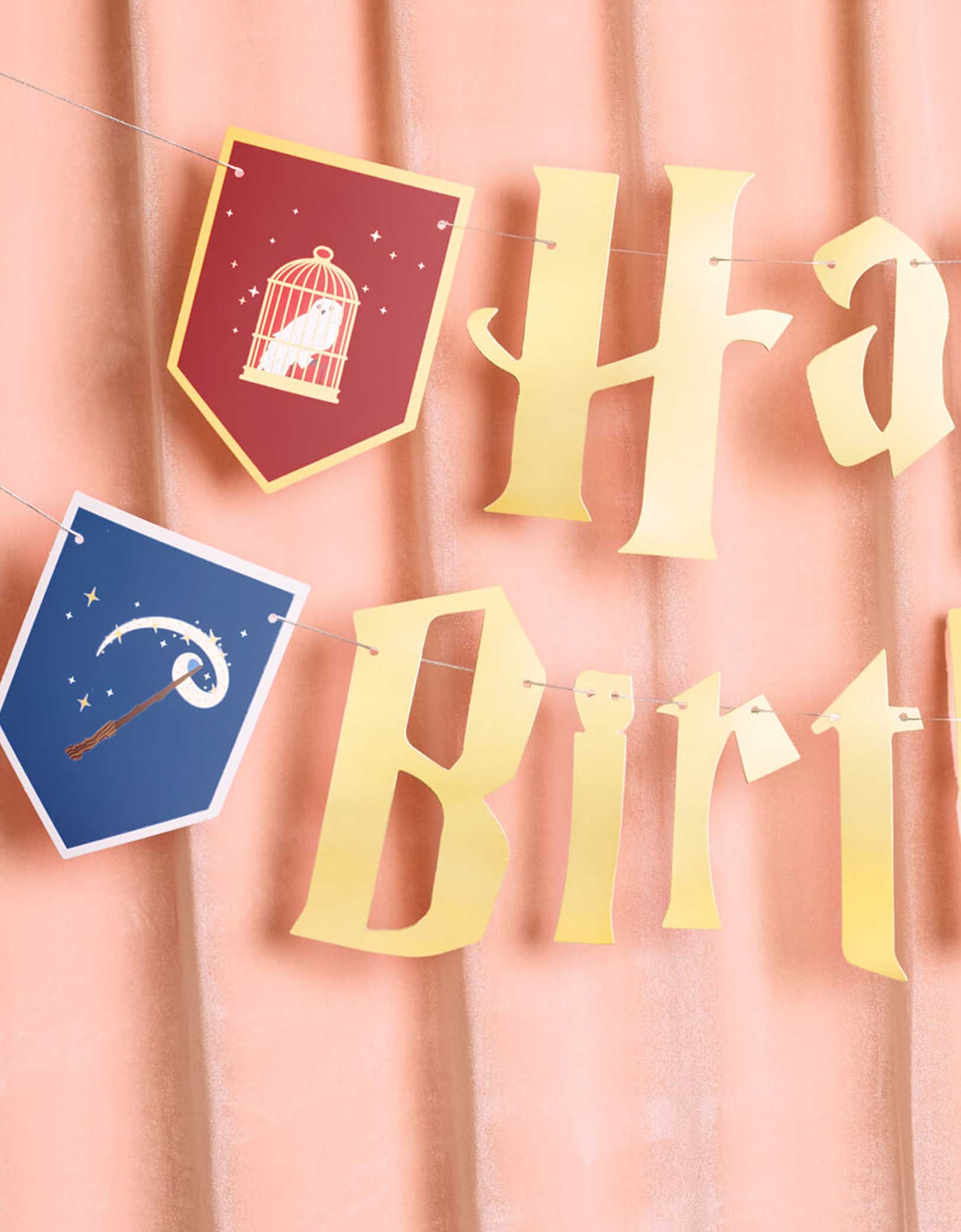 Momo Party's 5' Harry Potter themed Wizard Birthday Party Banner by Xo, Fetti. Inspired by the Hogwarts in Harry Potter, this party banner features "Happy Birthday" letters with red, green, blue and  yellow Harry Potter colored house crest shaped pendants with designs of an owl, a wand, a spell book, and a cauldron.  It's perfect for kid's Harry Potter themed birthday party!