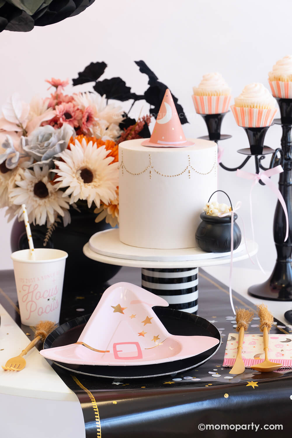 An adorable witch-themed pink Halloween party table setting featuring various pink Halloween party tableware from Momopartys including the pink witch hat shaped plate, Hocus Pocus small napkins, party cups and witch broom cutlery set on the mystic table runner. On the table there's a pink fondant cake adorned with a pink witch hat as the topper and a mini cauldron tied with a pink ribbon. With the pink boho floral arrangement and pink cupcakes on the vintage black candle holder a spooky cute table is ready!
