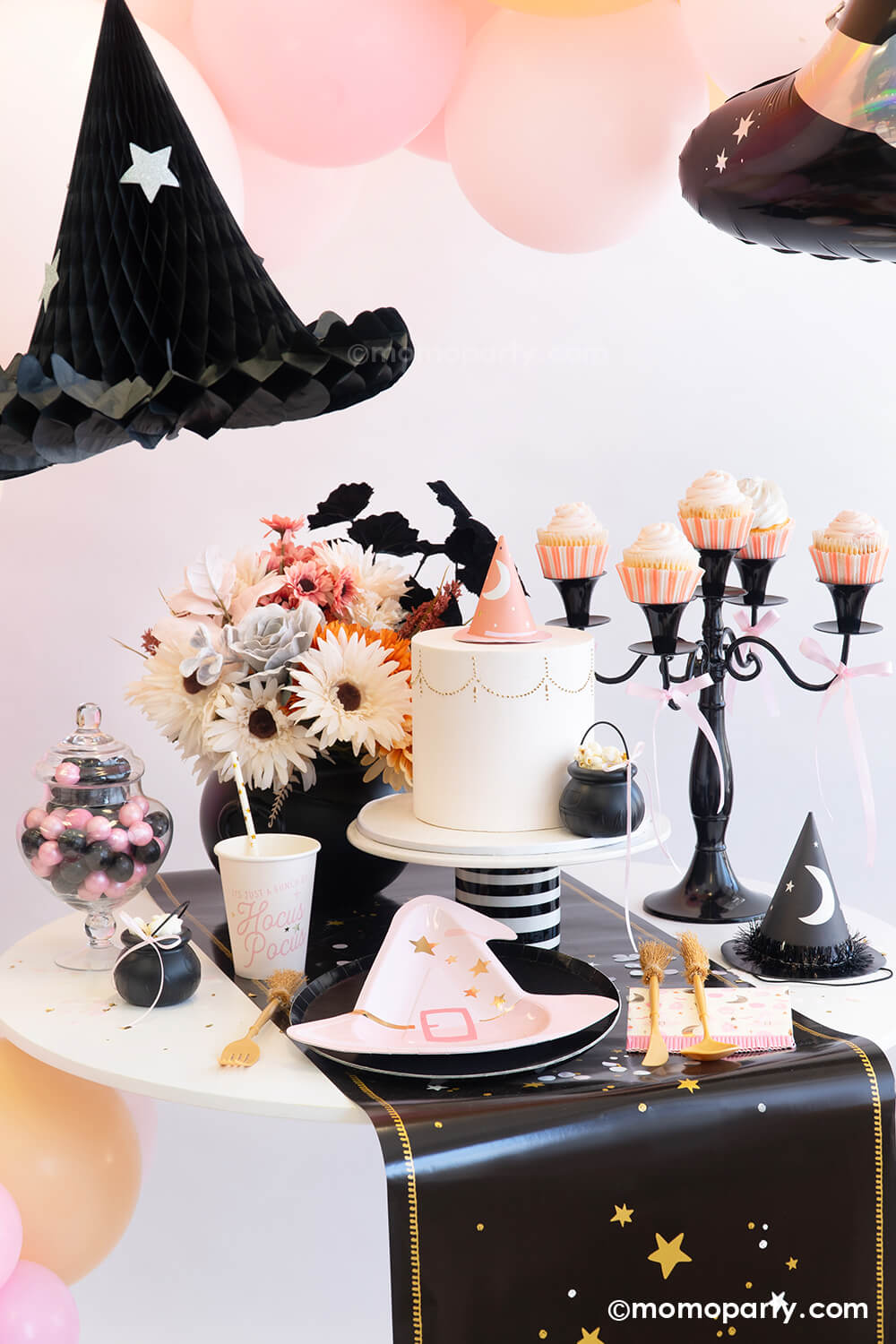 An adorable witch-themed pink Halloween party set up featuring various pink Halloween party supplies from Momopartys including the pink plates, cups, napkins and cutlery set. On the table there's a pink fondant cake adorned with a pink witch hat as the topper and a mini cauldron tied with a pink ribbon. On the table there are the pink boho floral arrangement, black candle holder and a gum bar jar. Above the table there's a pink balloon garland adorned with the witch hat honeycomb hanging decoration.