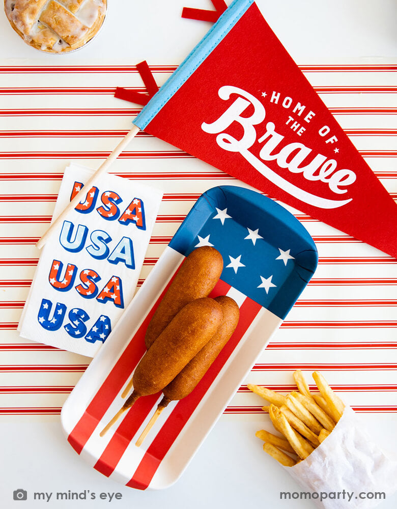 A festive patriotic themed party table featuring Momopartys's 15" Home of the Brave red felt pennant by My Mind's Eye, American flag shaped mini bamboo serving tray with summer snacks like corndogs and fries, next to it is a guest napkin with retro type of USA on it. With a matching red striped table runner, this makes a great inspo for a Memorial Day celebration or 4th of July Independence Day party.