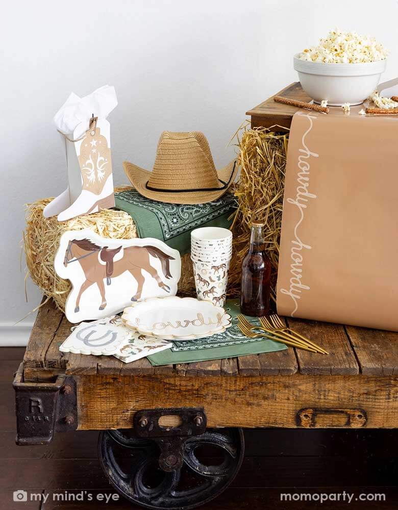 A cowboy themed party table featuring Momopartys's cowboy themed party supplies including horse shaped plates, cowboy patterned cups, napkins, table runner and cowboy boot shaped treat boxes. On the table there's a cowboy hat, straw bale, and a green bandana as decoration, perfect for a Western rodeo themed celebration, be a First Rodeo boy's first birthday party or a fun cowboy bash!