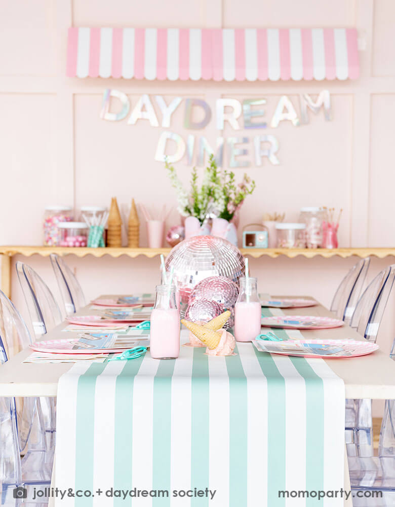An adorable kid's ice cream themed birthday party tablescape featuring Momopartys's ice cream truck shaped plate, the pink checkered plate, the ice cream icon napkin and pink wooden cutlery on a mint striped table runner. Behind the table there's a sundae station with sprinkle toppings, ice cream cones, adorned with a retro pink striped awning as a small ice cream shop, making this a cute, fun and festive ice cream party set up for kids birthday celebration.