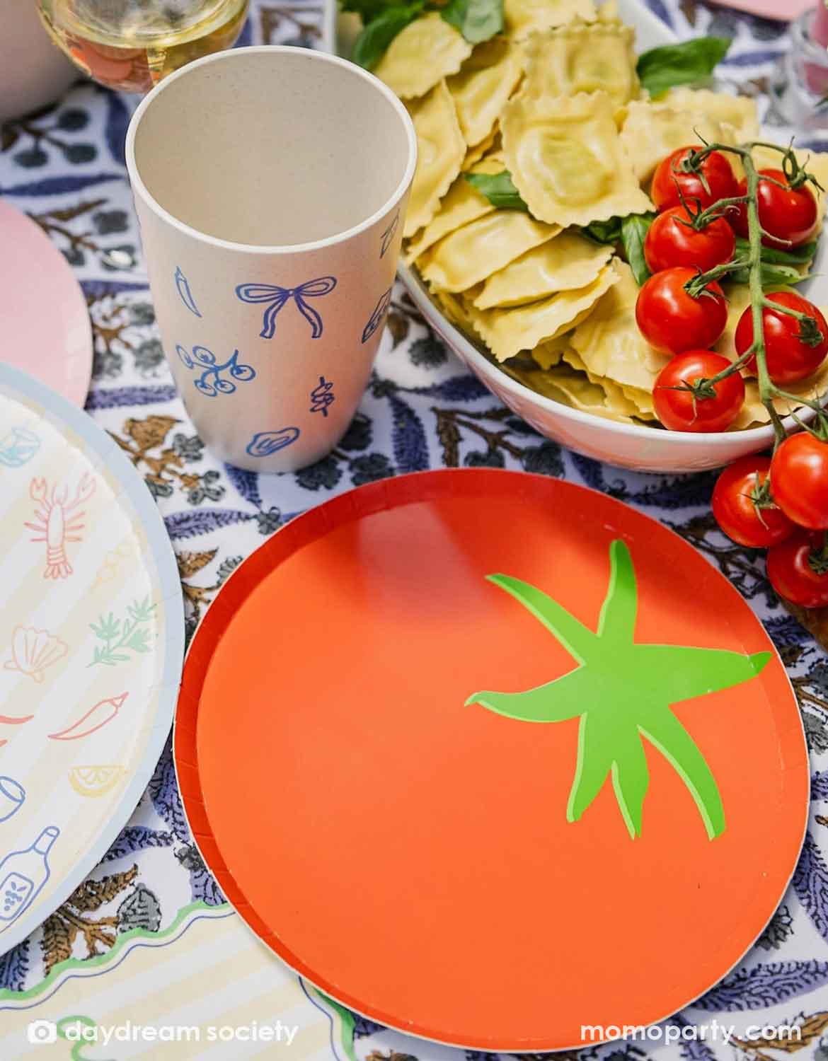 A festive Italian-inspired party table setting with Momopartys's 8" tomato shaped plate surrounded by a bowl of pastas and tomatoes with plates, cups and drinks around.