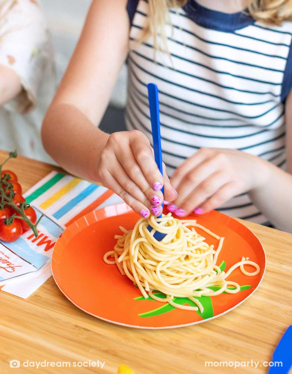 Child's hand with a blue plastic fork in spaghetti on Momopartys's tomato shaped plate with green leaves.