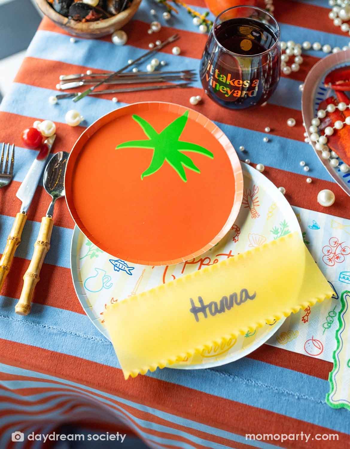 A festive Italian-inspired party table setting with Momopartys's 8" tomato shaped plate surrounded by a bowl of pastas and tomatoes with plates, cups and drinks around on a blue and red striped tablecloth.
