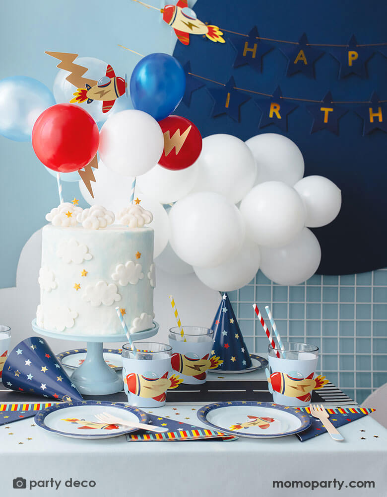 A kid's airplane birthday party decoration featuring Momopartys's plane themed party supplies and decorations by Party Deco, including 7" vintage airplane round paper plates, navy and star around large napkins and party hats, vintage airplane cup sleeves wrapped around party cups with red and blue striped paper straws. In the middle of the table is a birthday cake topped with airplane toppers and colorful mini balloons. In the back there's a blue backdrop with happy birthday banner hung on it. 