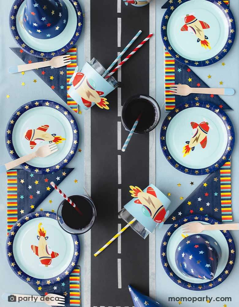 A top view shot of kid's airplane themed party table featuring Momopartys's 7" vintage airplane round paper plates, navy and star around large napkins, vintage airplane cup sleeves wrapped around party cups with red and blue striped paper straws. In the middle of the table is airplane runway track,  making this an adorable inspo for kid's aviation themed birthday party table setting.