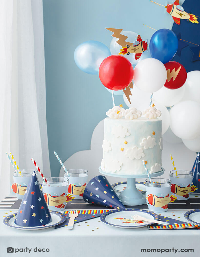 A kid's airplane birthday party decoration featuring Momopartys's plane themed party supplies and decorations by Party Deco, including 7" vintage airplane round paper plates, navy and star around large napkins and party hats, vintage airplane cup sleeves wrapped around party cups with red and blue striped paper straws. In the middle of the table is a birthday cake with cloud designs topped with airplane toppers and colorful mini balloons. Makes this an adorable inspo for kid's aviation birthday party.