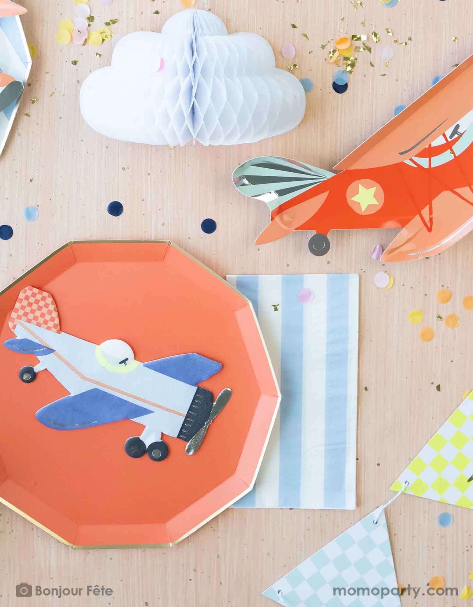 A kid's airplane themed birthday party tablescape featuring Momopartys's plane themed party supplies including Meri Meri's vintage airplane shaped napkin, plate and party garland pennants. A cabana inspired striped sky blue guest napkin is paired with the red decagon plate with gold foil edge. On the table there's an adorable honeycomb cloud and some festive confetti, making this a perfect inspiration for a modern and cute table setting idea for a boy's birthday party.