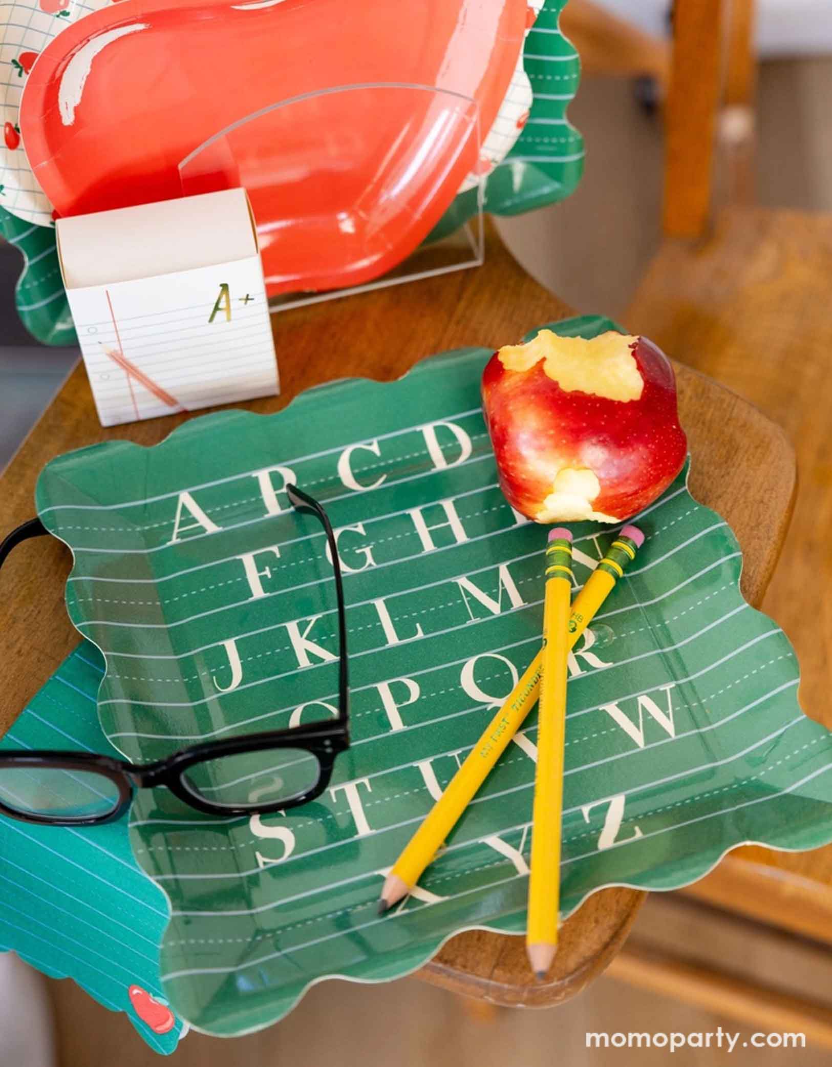 A back to school themed party table placeset featuring various school themed party supplies from Momopartys including the alphabet green large plate with matching Green Ruled Paper Dinner Napkins, along with apple shaped side plate, apple patterned scalloped plate and a notebook shaped treat box. On the alphabet plate there's a half-eaten apple and pair of glasses all on a wooden school chair.