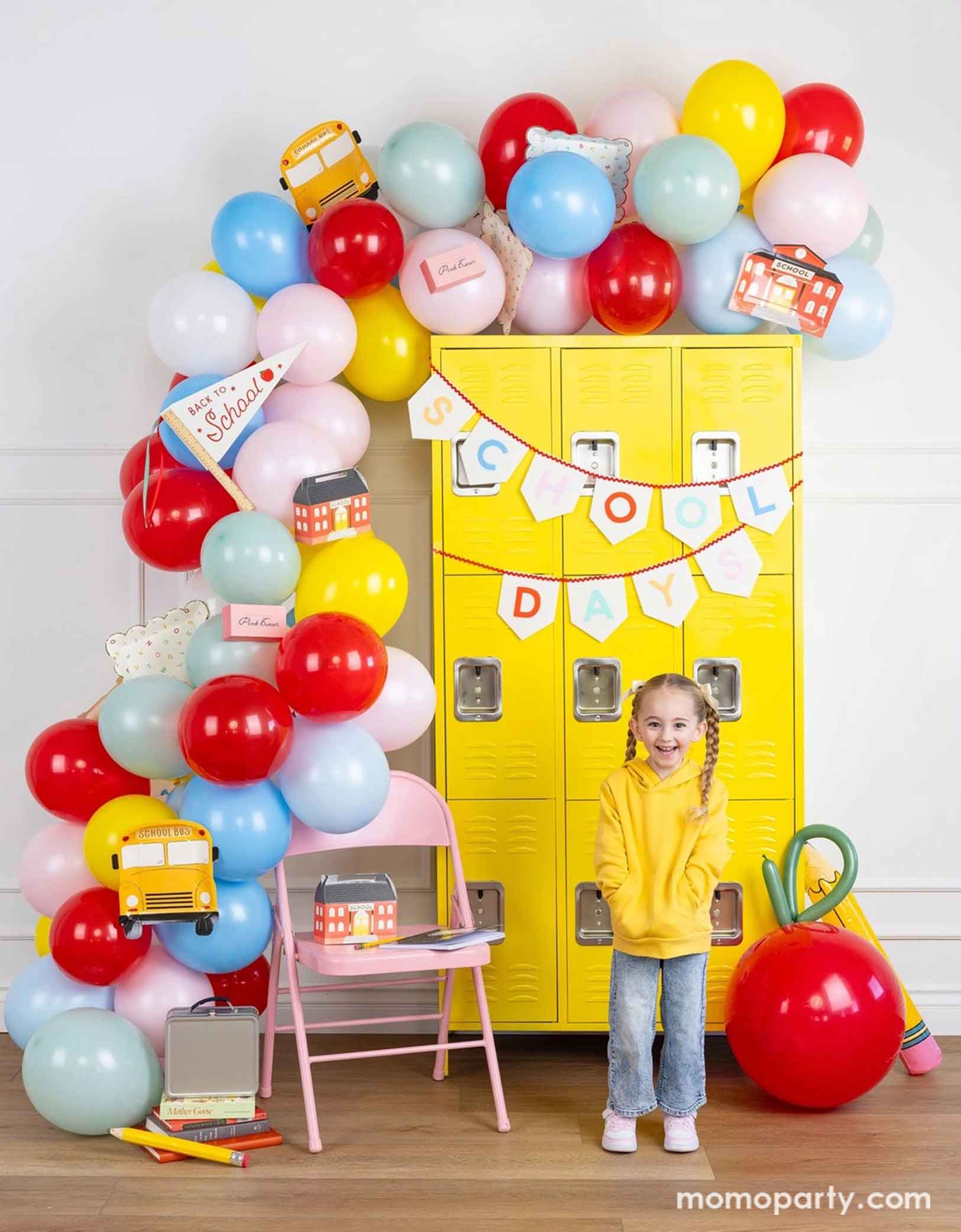 A girl standing in front of a first day of school celebration decoration set up featuring various school themed party decors from Momopartys including school house shaped treat houses and notebook plates on the chair, next to it is a festive colorful balloon garland adorned with school bus shaped plates, eraser shaped treat boxes, first day of school felt party pennant, and alphabet dinner plates. There's a yellow locker wall with SCHOOL DAY felt banner and a jumbo apple and pencil balloons.