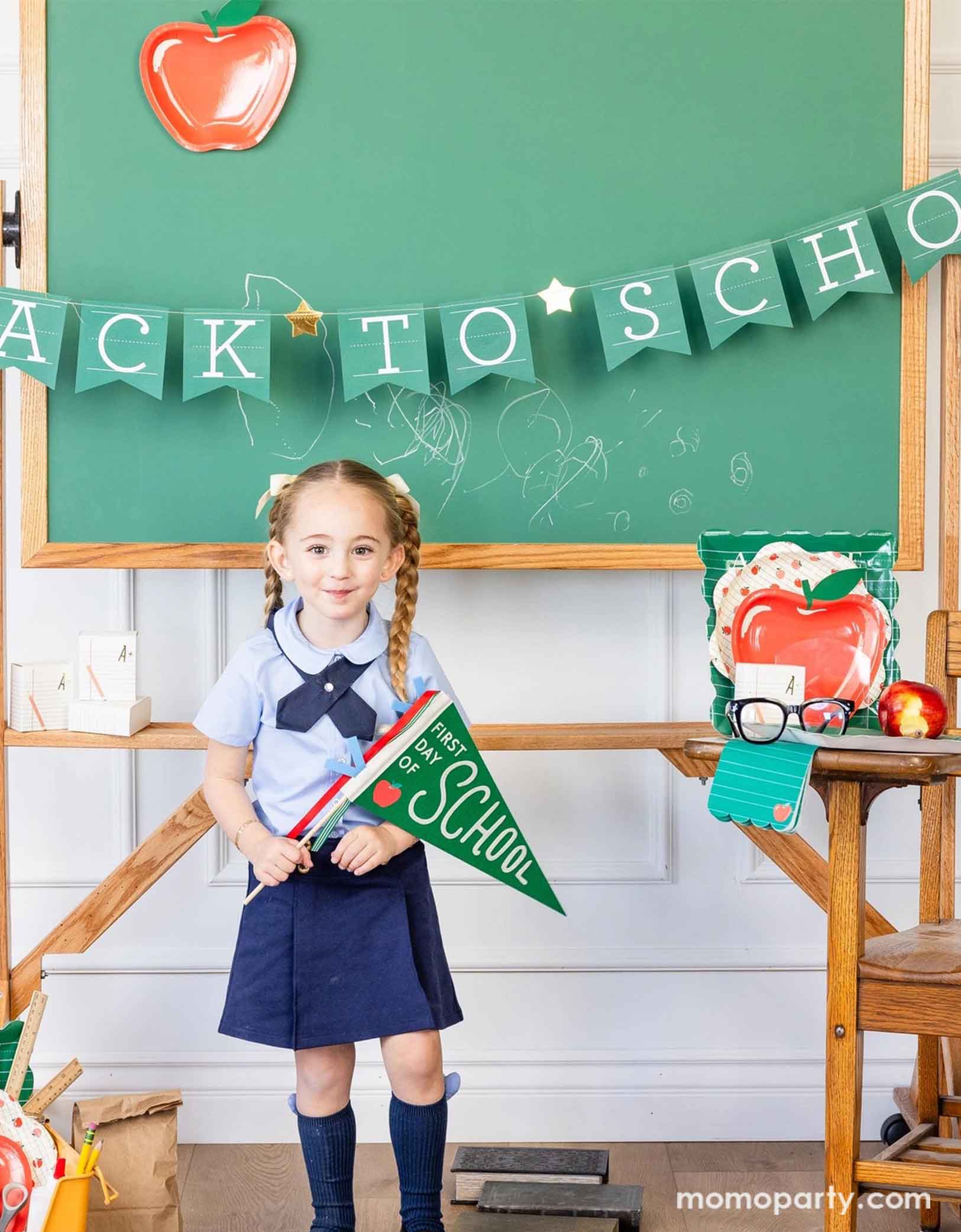 A school-aged girl in her blue uniform holding Momopartys's green first day of school apple felt pennant standing in a classroom setting decorated with a bunch of school themed party supplies from Momopartys including apple shaped plates, alphabet dinner plates and matching napkins. Behind her is a classroom chalkboard with back to school party banner hung across with an apple shaped plate - an adorable and cute set up to celebrate kid's back to school season!