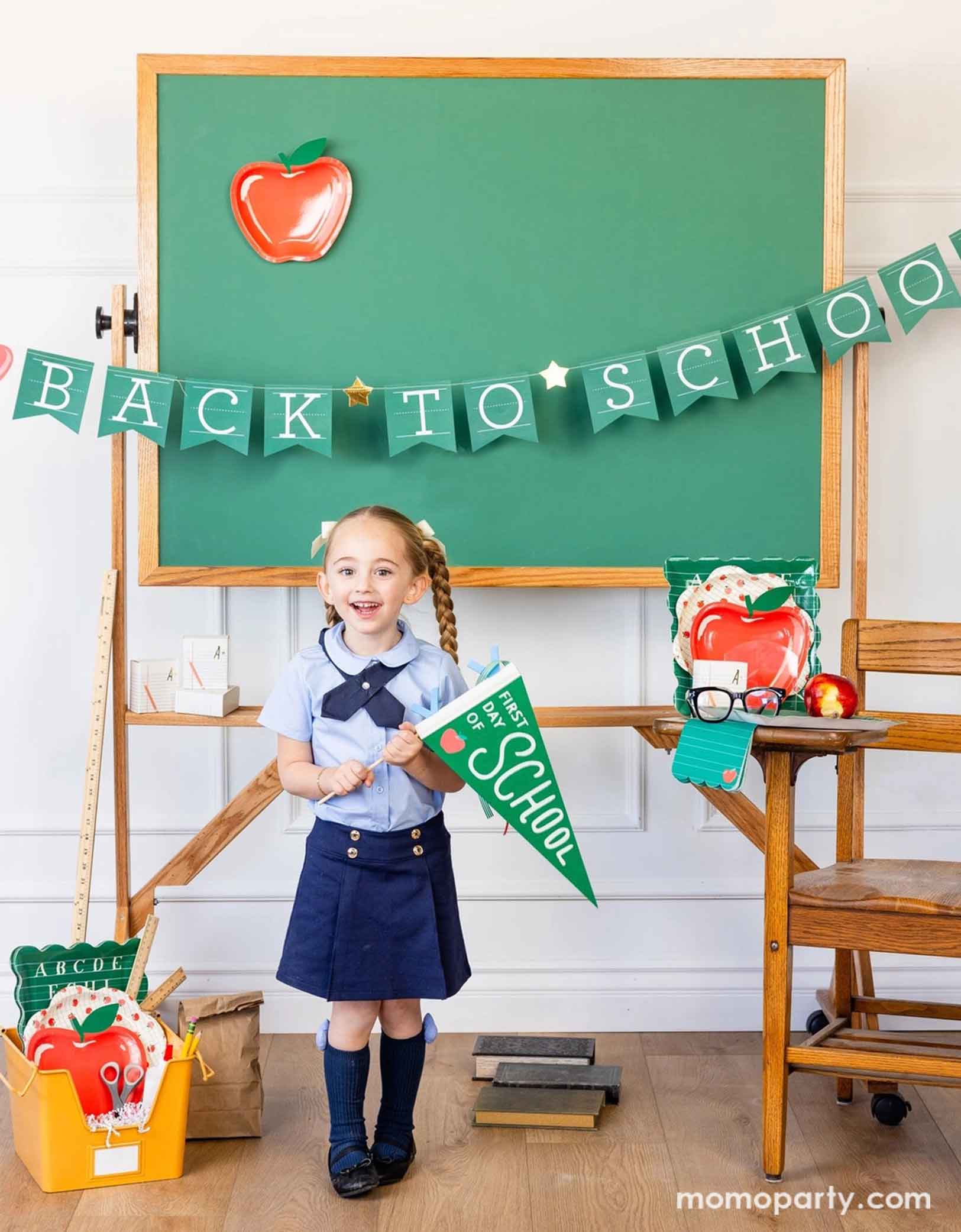 A school-aged girl in her blue uniform holding Momopartys's green first day of school apple felt pennant standing in a classroom setting decorated with a bunch of school themed party supplies from Momopartys including apple shaped plates, alphabet dinner plates and matching napkins. Behind her is a classroom chalkboard with back to school party banner hung across with an apple shaped plate - an adorable and cute set up to celebrate kid's back to school season!