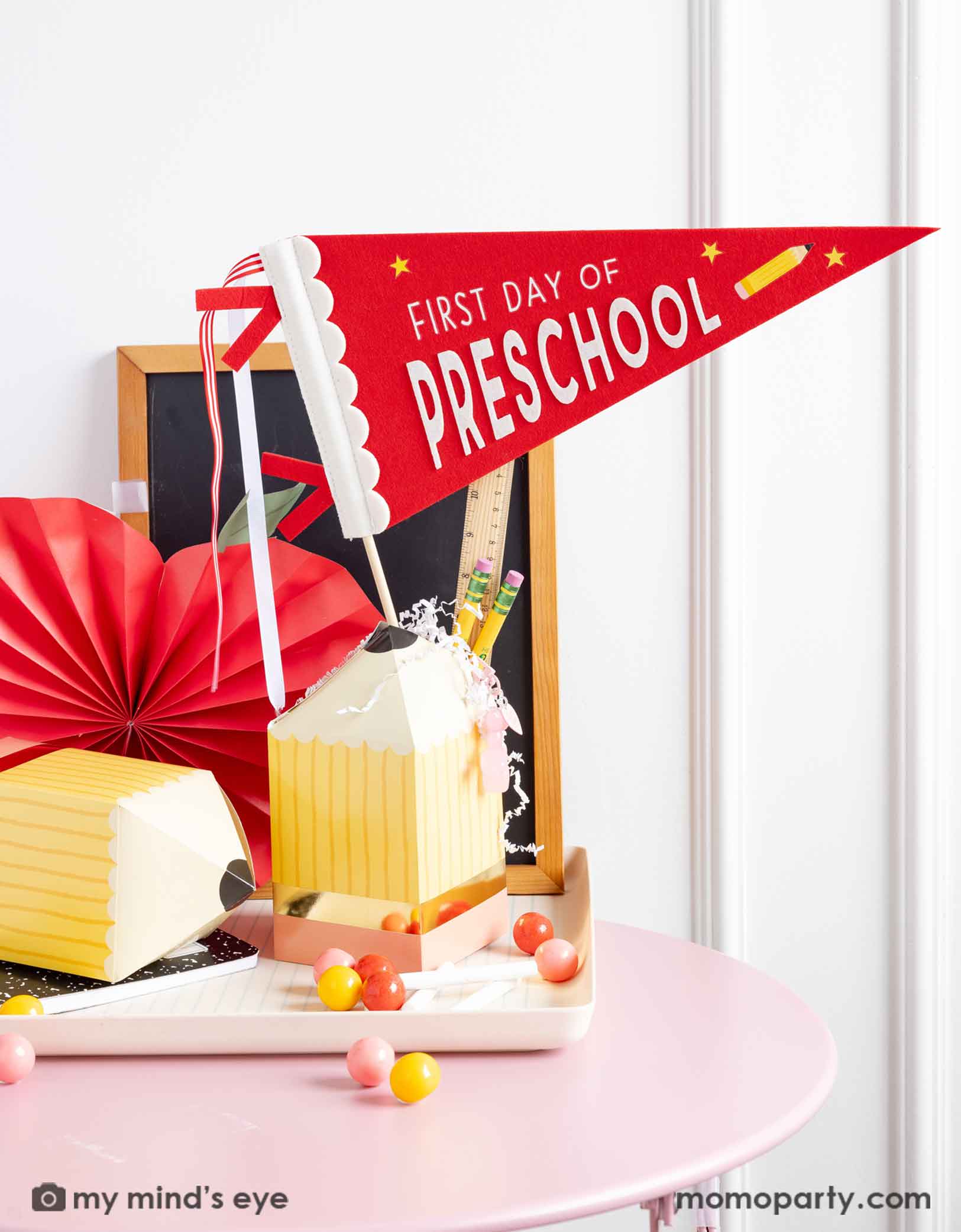 A festive back to school table set up decorated with various school themed party supplies from Momopartys including the red first day of preschool party pennant, pencil shaped treat boxes, notebook shaped bamboo party tray and apple shaped party fan. With a mini chalkboard in the back and some school supplies like pencils, rulers and some gumballs on the table, it creates a cute and modern look for a first day of school celebration.