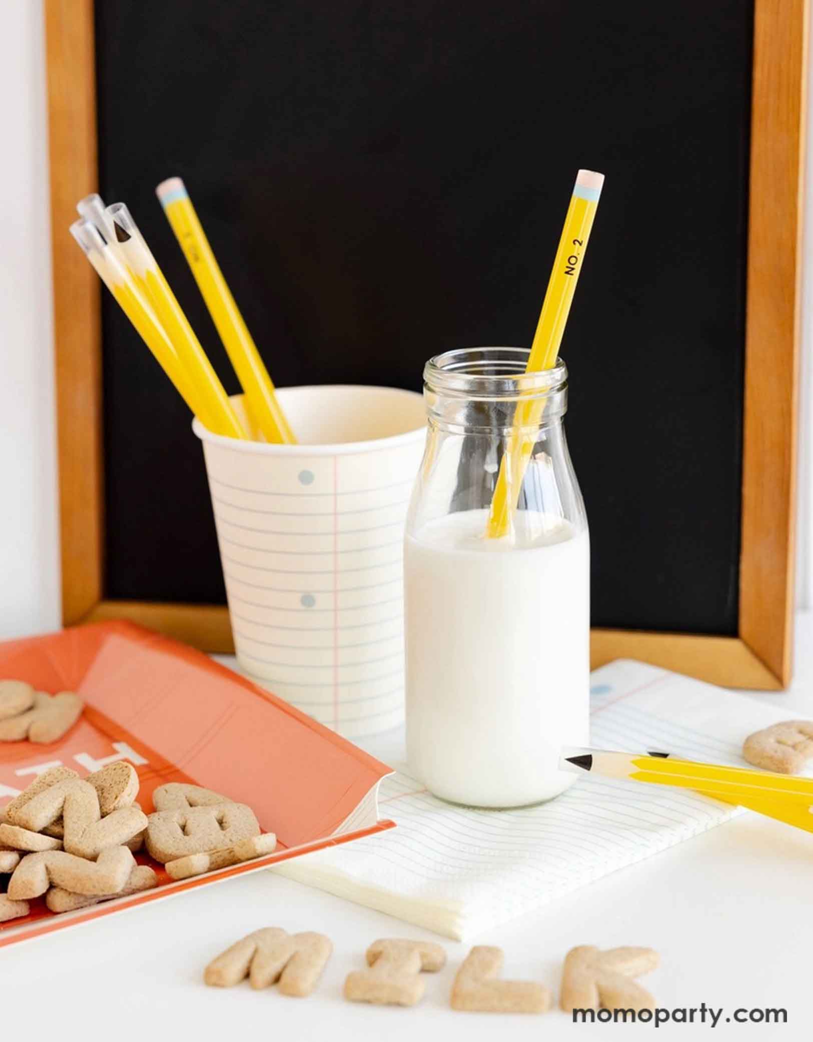 A clean and modern breakfast table featuring Momopartys's school themed party supplies including the 8oz notebook party cup with pencil shaped reusable straws, a stack of notebook paper guest napkins and a classic milk bottle filled with milk. On the table there are some alphabet cookies spelling out MILK and a small chalkboard in the back, making it a perfect info for first day of school breakfast set up for kids!