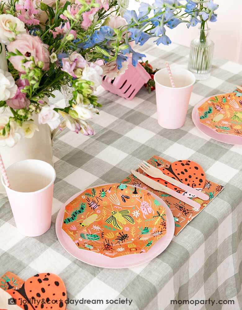 A cute kid's bug insect themed party table set up featuring Momopartys's backyard bug patterned plates, napkins paired with pink party cups and plates. With an olive green gingham tablecloth under and a beautiful flower bouquet, this makes a perfect inspiration for a modern take of a kid's bug themed celebration.