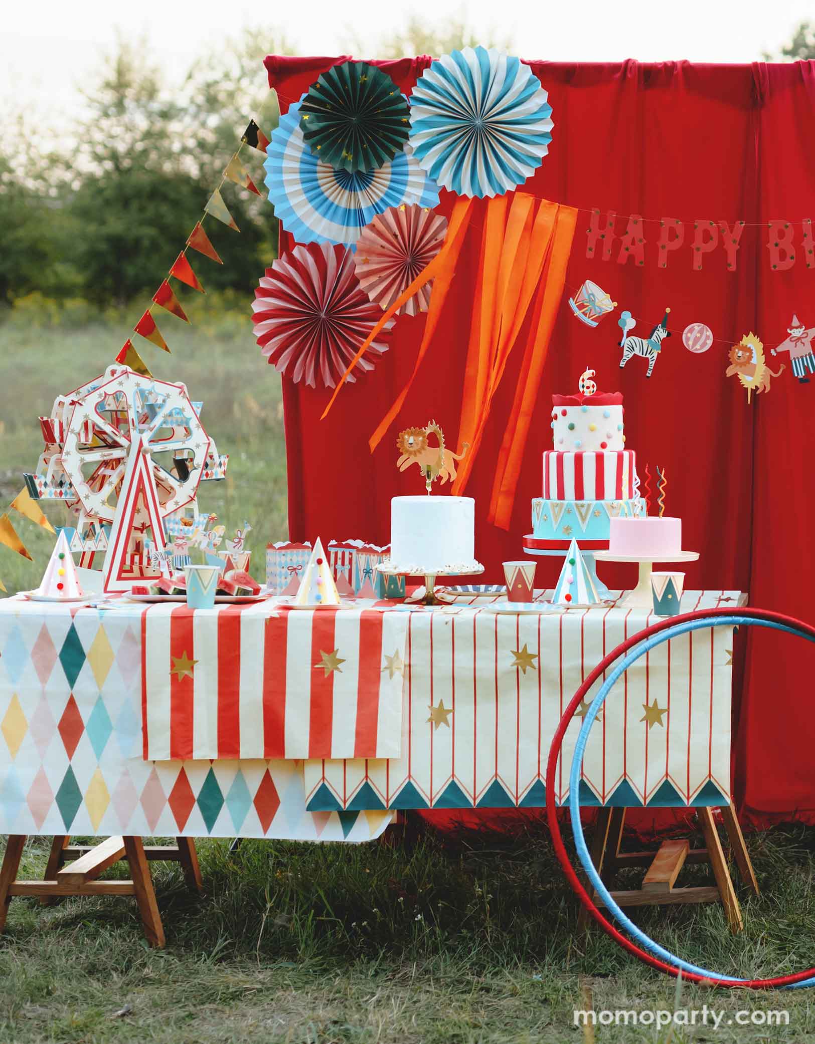 An adorable circus & carnival themed birthday party set up features various circus themed party supplies from Momopartys, including a stunning ferris wheel cupcake stand centerpiece with circus-inspired design, party plates, hats, cups, treat boxes and multiple birthday cakes on a carnival themed tablecloth. In the back there's a red curtain as a party backdrop decorated with circus birthday banner with circus animal party pennants and colorful party fans. A perfect inspo for a fun circus party for kids.
