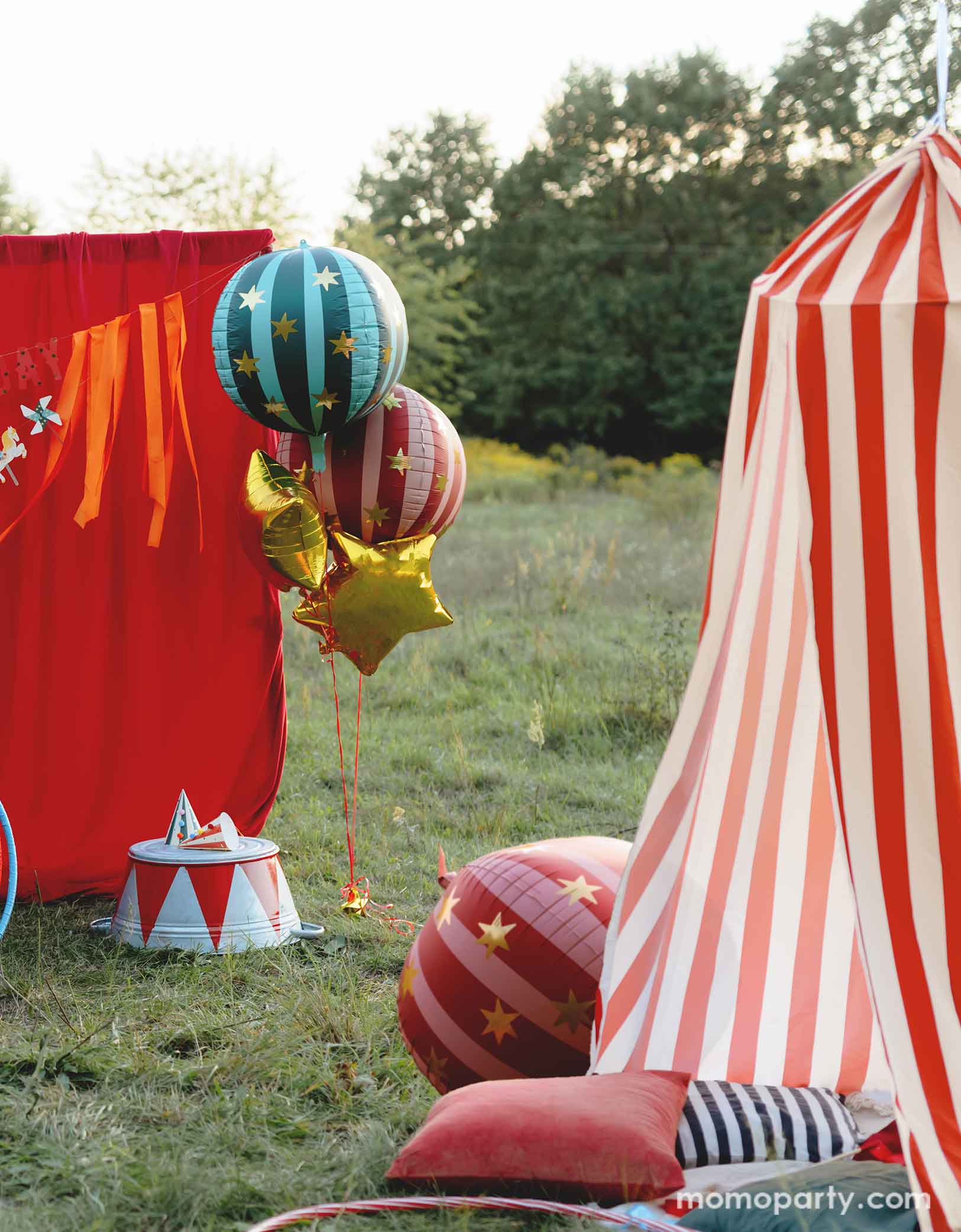 A vintage circus themed party set up in the park featuring various carnival circus themed party decorations from Momopartys including the red striped globe foil balloon, the green striped globe foil balloon and gold star foil balloons floating next to the red curtain party backdrop. In the front there's a red and white striped circus themed play tent for kids with some colorful balloons and a red striped globe foil balloon next to it. Making this a whimsical and playful setting for a fun kid's circus party.