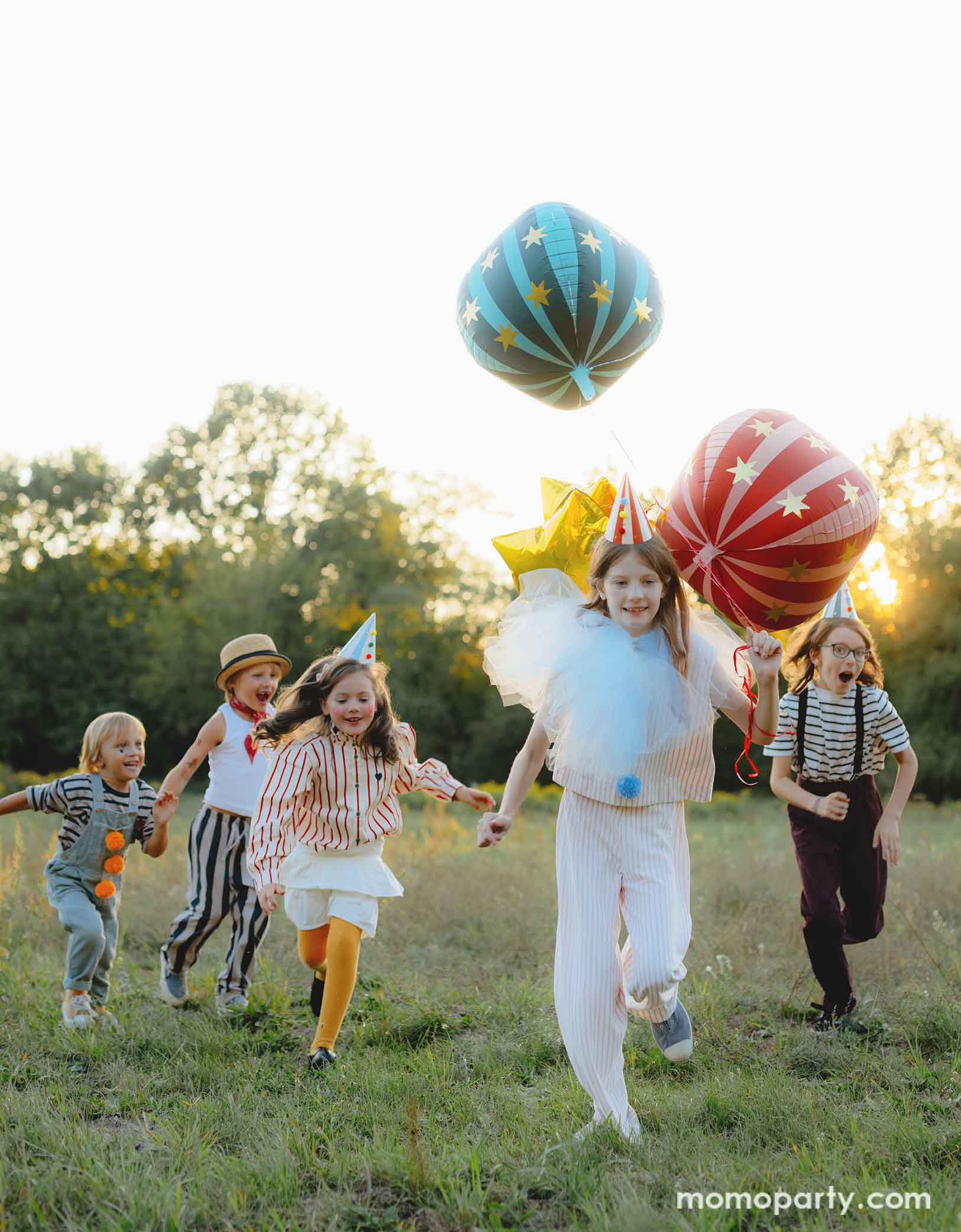 A bunch of kids dressed in quirky vintage circus inspired clothing holding Momopartys's circus themed party balloons running towards the camera. The foil balloons floating in the air include a red globe foil balloon, a green striped globe foil balloon and some gold star foil balloons, perfect decorations for a fun kid's circus/carnival themed celebration.