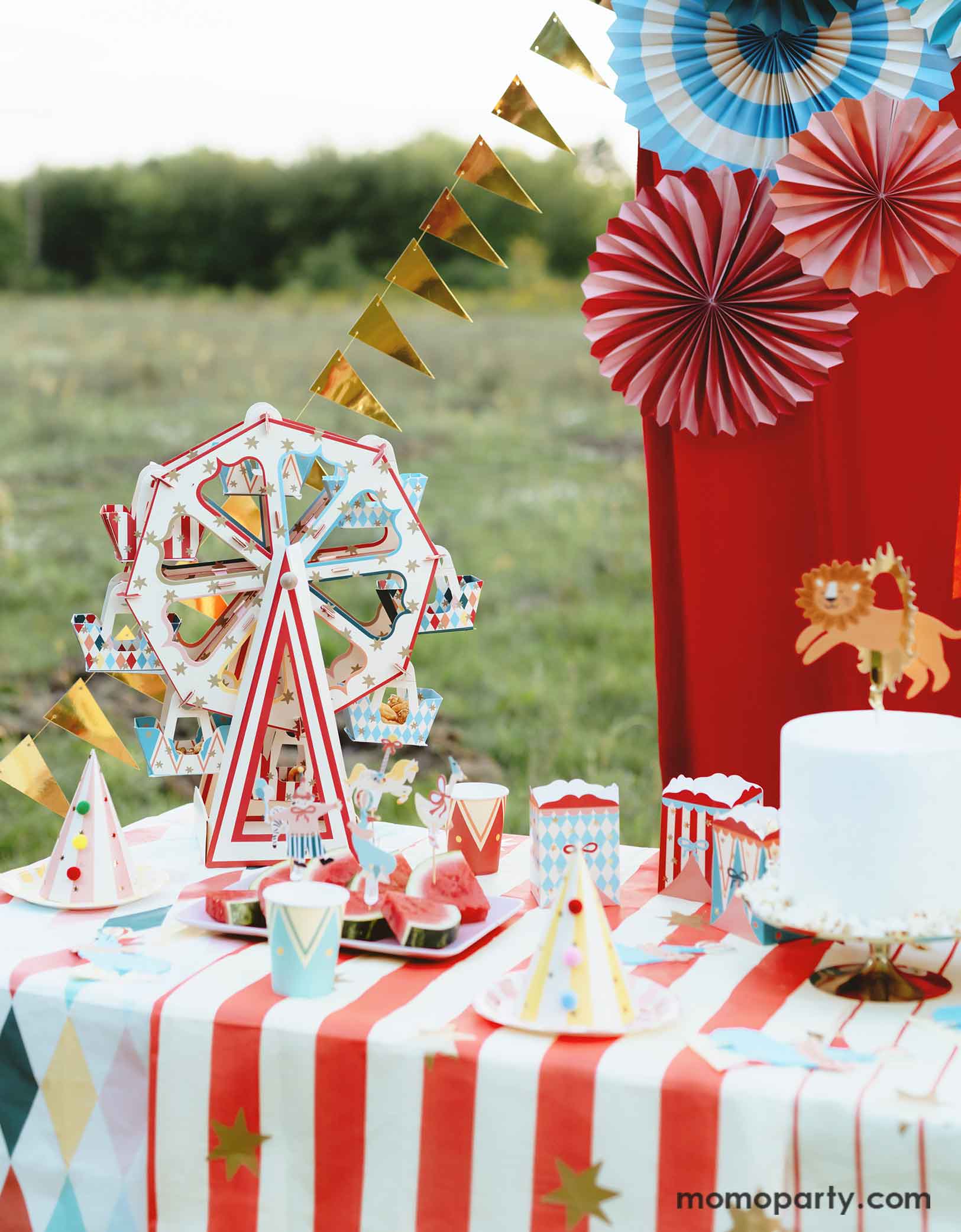 An adorable circus & carnival themed birthday party table set up and decorations feature various circus themed party supplies from Momopartys, including a stunning ferris wheel treat stand centerpiece with its circus-inspired design is loaded with cupcakes, party plates, hats, cups, treat boxes and a birthday cake on a carnival themed tablecloth. In the back there's a red stage curtain as a party backdrop decorated with circus themed birthday banner with circus animal party pennants and colorful party fans.