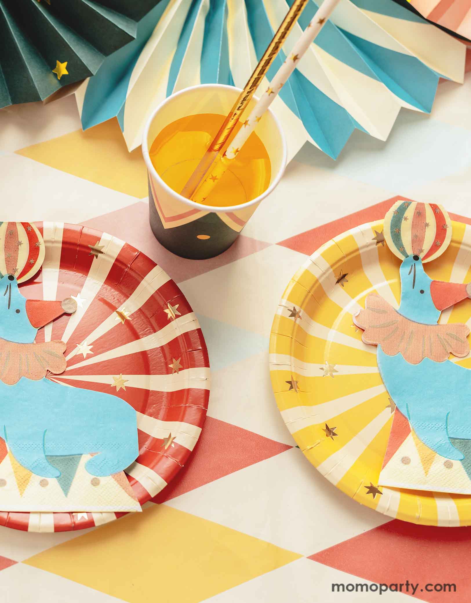 A carnival/circus themed part table featuring Momopartys's circus themed party tableware including a red and yellow striped paper plates and sea lion playing with a ball shaped napkins, with matching party cups with gold foil star paper straws and some festive party fans on a circus themed striped table cover - making this great table setting idea for a fun and festive kid's carnival themed birthday celebration!