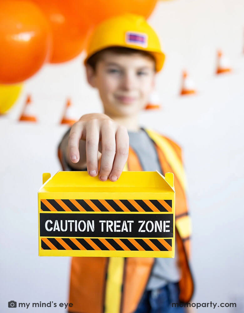 A school aged boy in his construction costume and hard hat holding Momopartys's toolbox shaped treat box by My Mind's Eye standing in front of a festive construction themed party decoration which features a balloon garland in black, orange and yellow and a construction cone banner hung over on the wall - a perfect decoration idea for kid's construction builder themed birthday celebration party.
