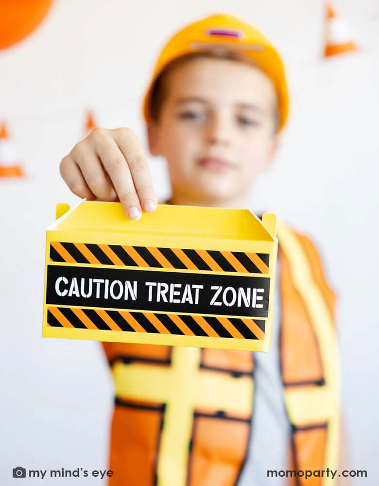 A school aged boy in his construction costume and hard hat holding Momopartys's toolbox shaped treat box by My Mind's Eye standing in front of a festive construction themed party decoration which features a balloon garland in black, orange and yellow and a construction cone banner hung over on the wall - a perfect decoration idea for kid's construction builder themed birthday celebration party.