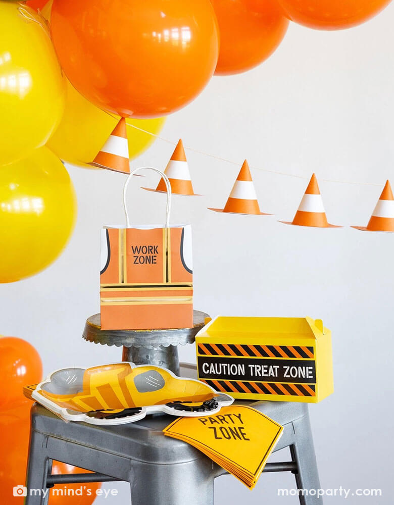 A festive kid's construction themed birthday party decoration set up featuring various construction party supplies and decorations from Momopartys including dumper truck shaped plates, party zone napkins, work zone vest shaped party bags, construction toolbox shaped treat boxes by My Mind's Eye. In the back there's a balloon garland in classic construction colors of orange and yellow, adorned with a construction cone paper banner, making this a perfect inspo for boy's construction birthday party!