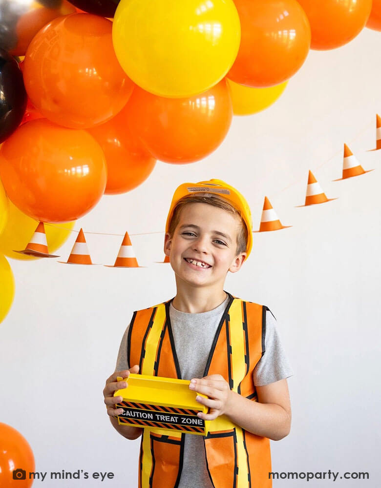 A school aged boy in his construction costume and hard hat holding a toolbox shaped treat box standing in front of a festive construction themed party decoration which features a balloon garland in black, orange and yellow and a construction cone banner hung over on the wall - a perfect decoration idea for kid's construction builder themed birthday celebration party. 