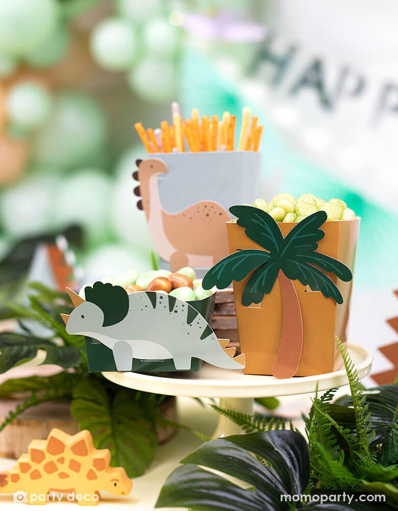 A kid's dinosaur themed party table features Momopartys's dinosaur snack boxes with cute dinosaur and palm tree designs in soft colors on the side. Around the table are some tropical plants decorated, creating a pre historical vibe. In the back the are some mint green party balloons and Happy Birthday banner.