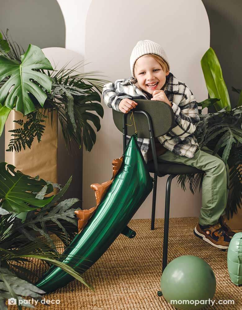 A boy dressed in green outfit sitting on a green metal chair decorated with Momopartys's 29" x 10" dark green dinosaur tail shaped foil balloon. The room is dressed with lots of tropical plants and sage green party balloons, setting a great prehistoric scene for a kid's dinosaur themed party celebration!