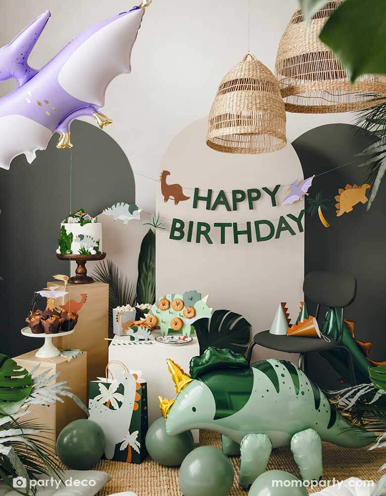 An adorable kid's dinosaur themed party set up featuring Momopartys's dinosaur themed party supplies, decorations and balloons by Party Deco including a mint Triceratops shaped foil balloon, a lilac Pterodactyl shaped foil balloon, sage green party balloons, various dinosaur cake toppers, dinosaur spikes party hats, and a dinosaur happy birthday banner hung on the party backdrop wall. All makes a cute and modern inspo for kid's dinosaur themed birthday party.
