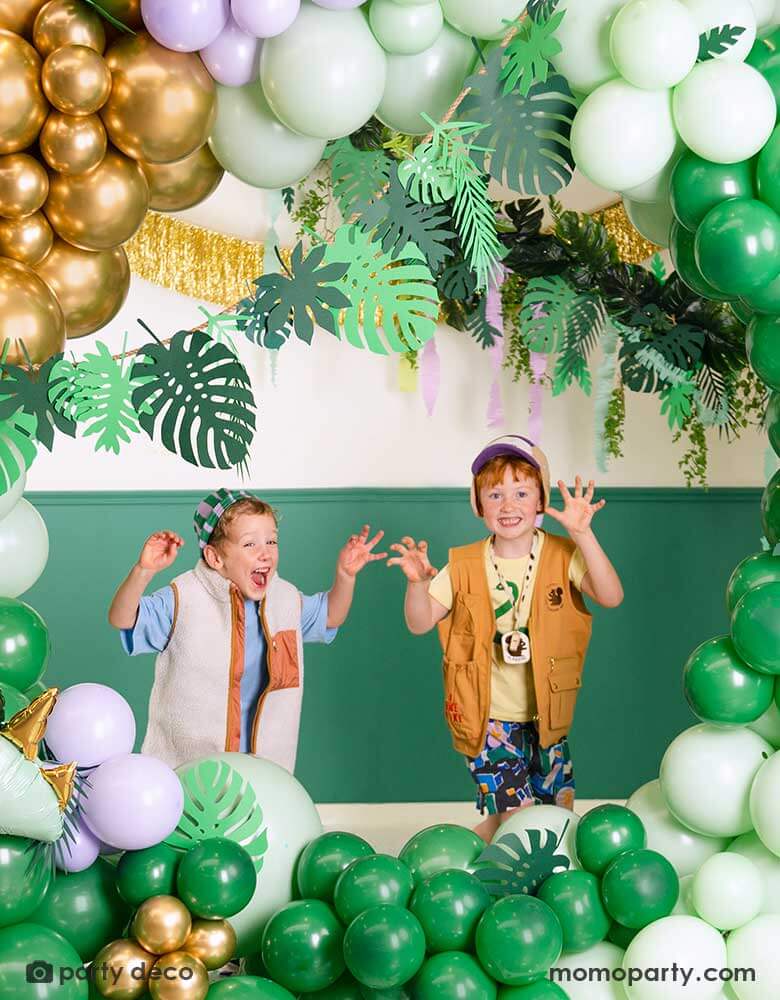 Two school aged boys in a tropical dinosaur themed balloon decoration set up featuring balloon garland in different shades of green, lilac and chrome gold. The organic balloon garland is adorned with Momopartys's tropical leaves paper decorations by Party Deco. Along with the gold tinsel fringe garland, it creates an amazing prehistorical vibe, a perfect inspo for kid's dinosaur themed birthday party or a tropical themed bash in summer!
