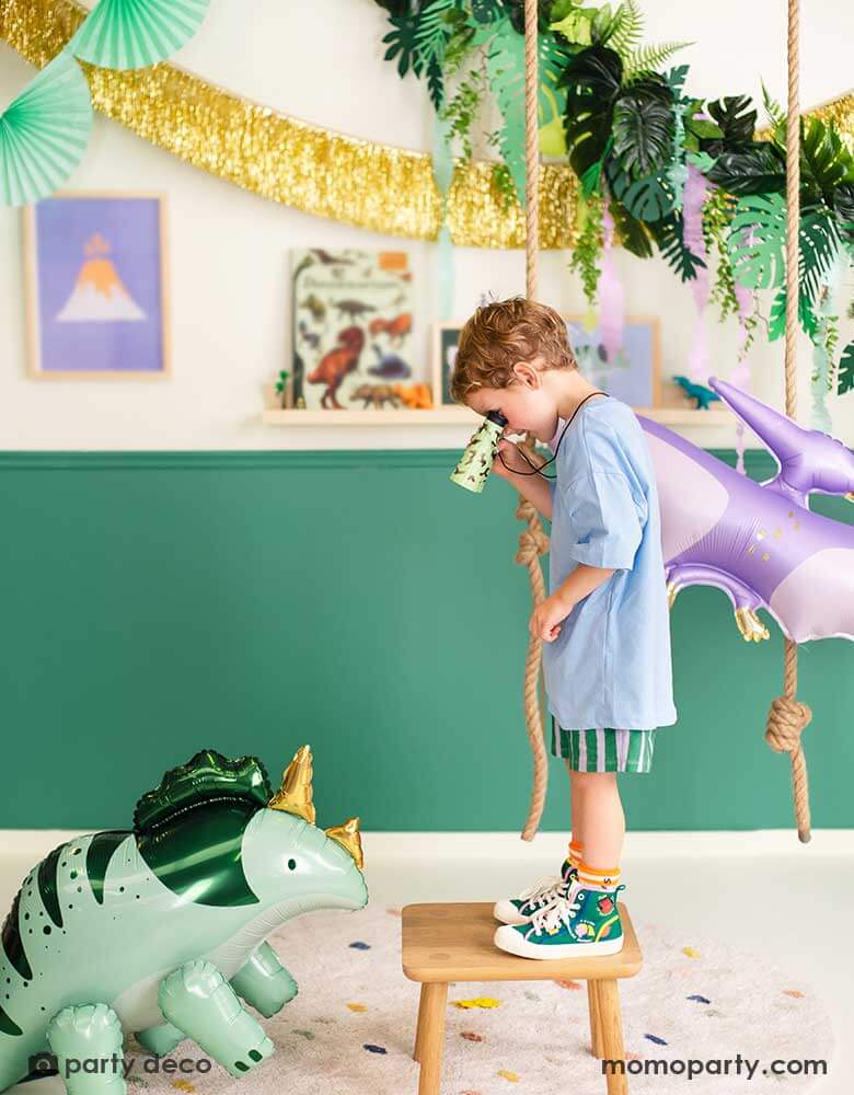 A boy is watching Momopartys's Triceratops shaped foil balloon through his binoculars. He's in a room decorated with prehistorical  themed decoration including a lilac PTERODACTYL shaped balloon, a gold tinsel fringe garland, a tropical leaves garland from Momopartys.